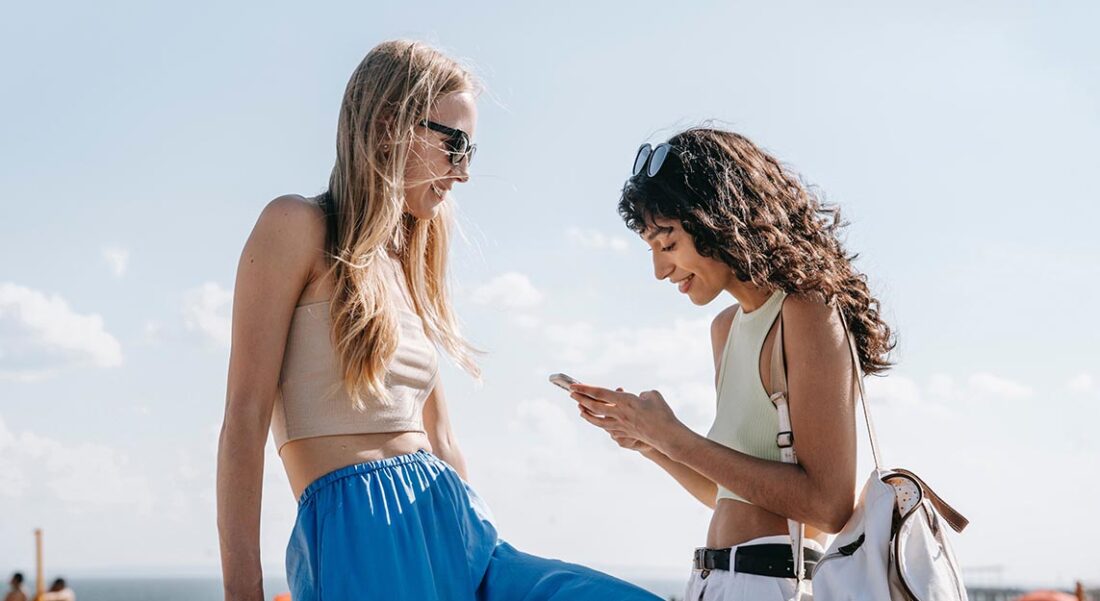 two young women outdoors with sunglasses; one is looking down at her phone